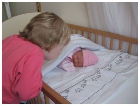 In een ledikantje ligt een pasgeboren  baby met een roze mutsje. Een blonde peuter met een roze trui leunt over de rand van het bedje. 