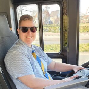 Lizzy zit met een glimlach achter het stuur van een bus. Ze heeft kort haar, draagt een zonnebril, uniformblouse en stropdas. 